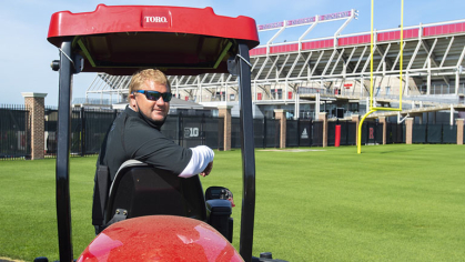 A Utilities Manager riding on a lawnmower looks back, satisfied, at the viewer. Beautiful and perfectly manicured turfgrass in Rutgers Stadium is behind him.