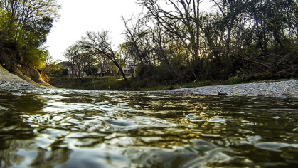 A view from right over the water of a babbling brook on a sunny day. The trees indicate a changing of the seasons is coming.