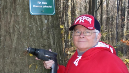 A Master Tree Stward holding an electric drill next to a tree that has a new sign on it that says 'Pin Oak (Quercus palustris)'.