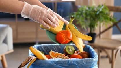 A person throwing vegetables and fruit into a garbage can.
